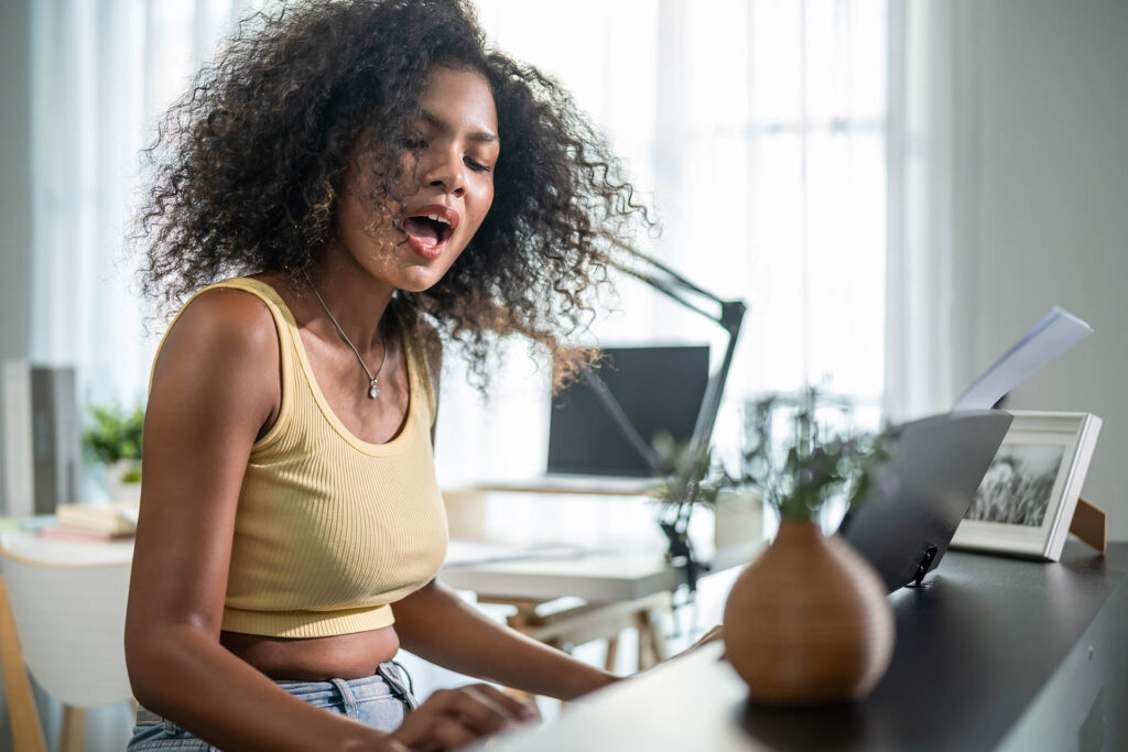 Woman singing and playing piano, comparing Online Voice Lessons vs In-Person coaching.